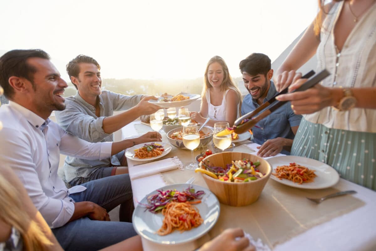 Friends and family Serving salad on a table at sunset. There is spaghetti Bolognese on some of the plates on the table. There are glasses of wine and other food on the table; everyone is having fun laughing and smiling